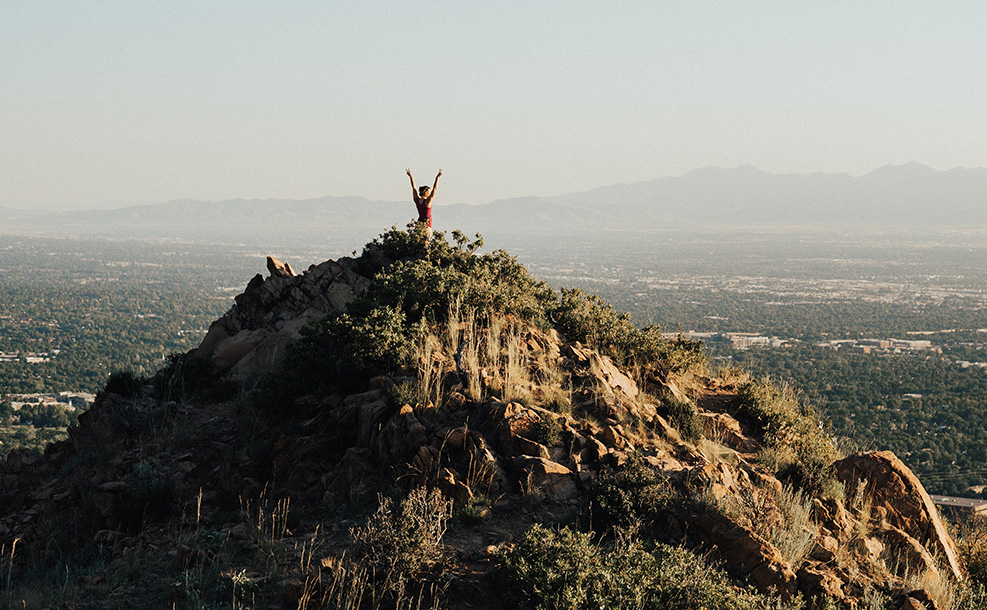 a person standing triumphantly, with their hands raised high, overlooking a forested valley